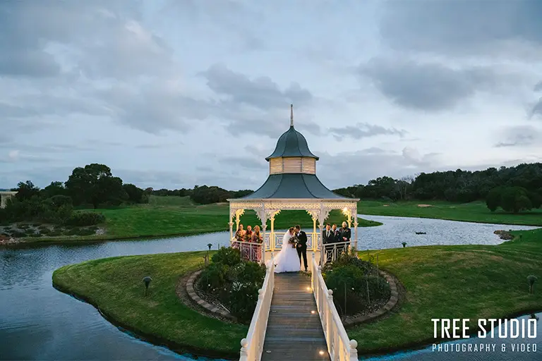 A bride and groom kiss inside a white gazebo decorated with lights at Eagle Ridge Golf Course in Victoria. The gazebo sits on a small island surrounded by water, with a wooden bridge leading across. Guests stand around the couple watching, while the golf course’s green fairways stretch out in the background under an evening sky.