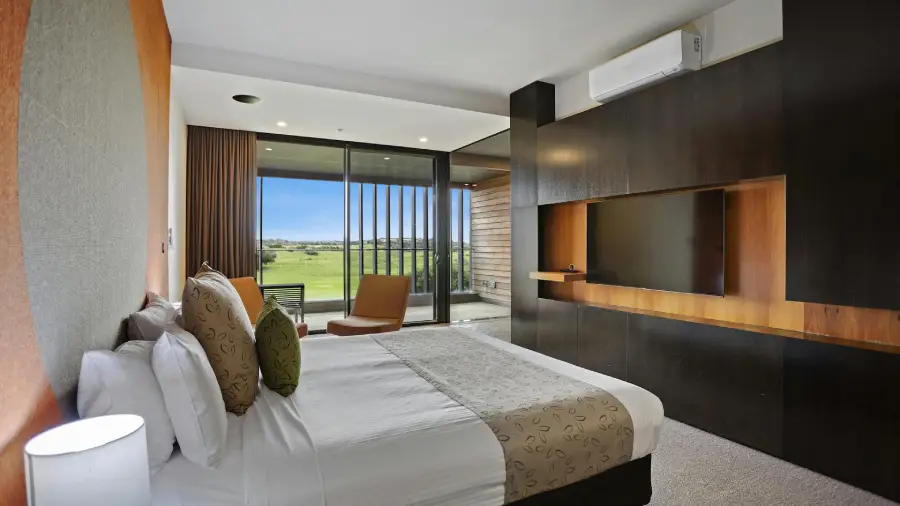 A modern hotel room at Peppers Resort, Moonah Links, featuring a large bed with crisp white linens and patterned cushions. Opposite the bed is a sleek, dark wood wall unit with a flat-screen TV and warm timber accents. Floor-to-ceiling sliding glass doors open to a private balcony with views of the green golf course beyond, bringing in natural light and a sense of openness. A tan armchair sits near the window, offering a cozy spot to relax and enjoy the scenery.
