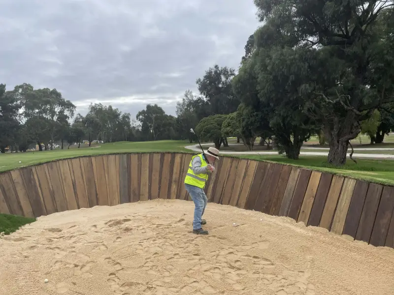 golfer in a high-visibility vest and hat takes a swing from the base of a steep timber-faced bunker at Freeway Golf Course in Melbourne. The bunker’s vertical wooden walls rise dramatically behind him, with trees and fairway visible in the background under a cloudy sky.