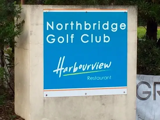 A blue sign mounted on a concrete base reads “Northbridge Golf Club – Harbourview Restaurant” in white and stylised script. The sign is surrounded by trees and foliage.