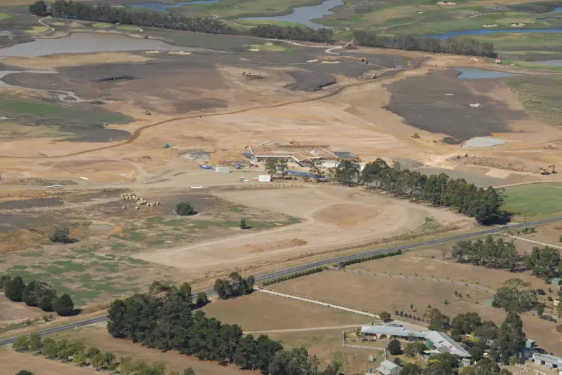 A 2007 aerial photo showing the early construction of Yering Meadows Golf Club in Victoria. The land is mostly bare earth and soil works, with patches of green grass beginning to appear. Earthmoving equipment and partially built structures are visible in the centre, while surrounding farmland and early water hazards take shape across the site.
