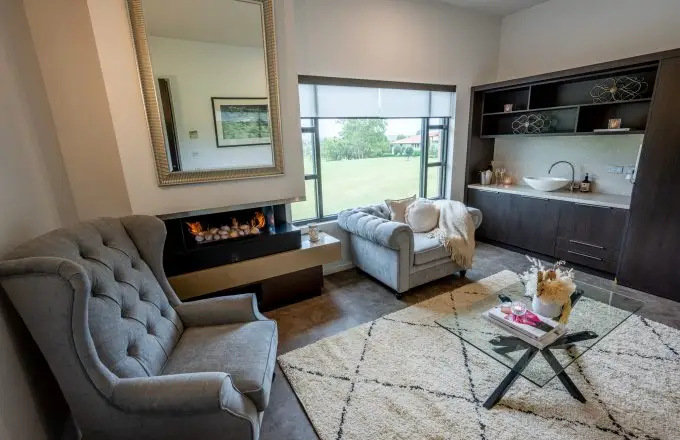 Cosy Château Élan lounge room at The Vintage Golf Club with grey armchairs, modern fireplace, glass coffee table, kitchenette, and window overlooking green lawns.